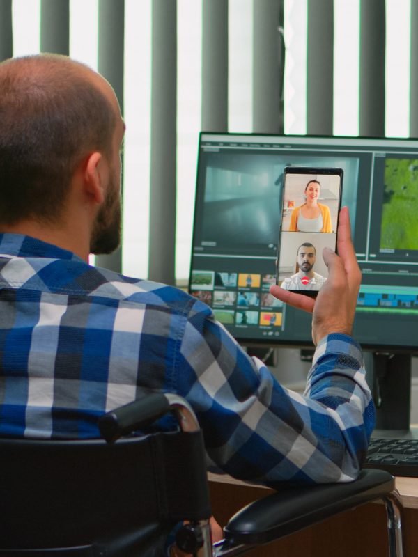 Handicapped videographer in wheelchair talking on webcam with cowerkers while editing a video project creating content in modern company office. Creator blogger working from photo studio.